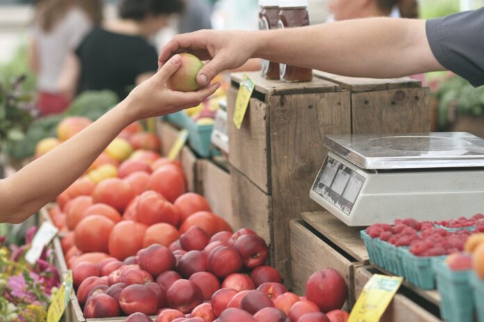 mercati all'ingrosso - in primo piano un banco da mercato di frutta dal quale spunta una mano che porge una mela ad un'altra mano presumibilmente di donna