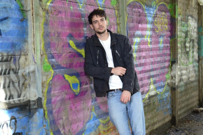wires - henry beckett in primo piano, indossa jeans azzutti, maglia bianca e giubbotto nero, ed è appoggiato ad un muro disegnato