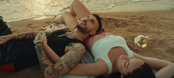 frame del video extasi un ragazzo sdraiato sulla spiaggia con accanto una ragazza guardano un tramonto