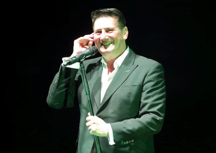 Sorridente si porta una mano allì'orecchio Tony Hadley indossa camicia bianca e giacca scura