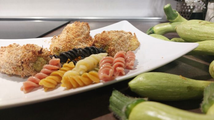 un piatto ben presentato con della pasta fusilli colorata, delle acciughe ripiene fritte e intorno al piatto (bianco e quadrato, ci sono delle zucchine