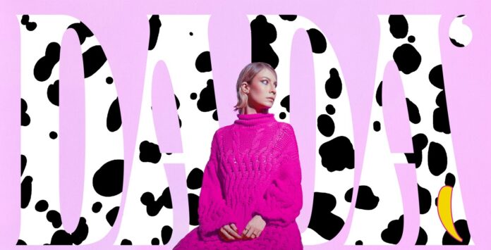 dadà di profilo indossa un maxi pullover color fucsia, alle spalle la scitta del nome a caratteri maculati