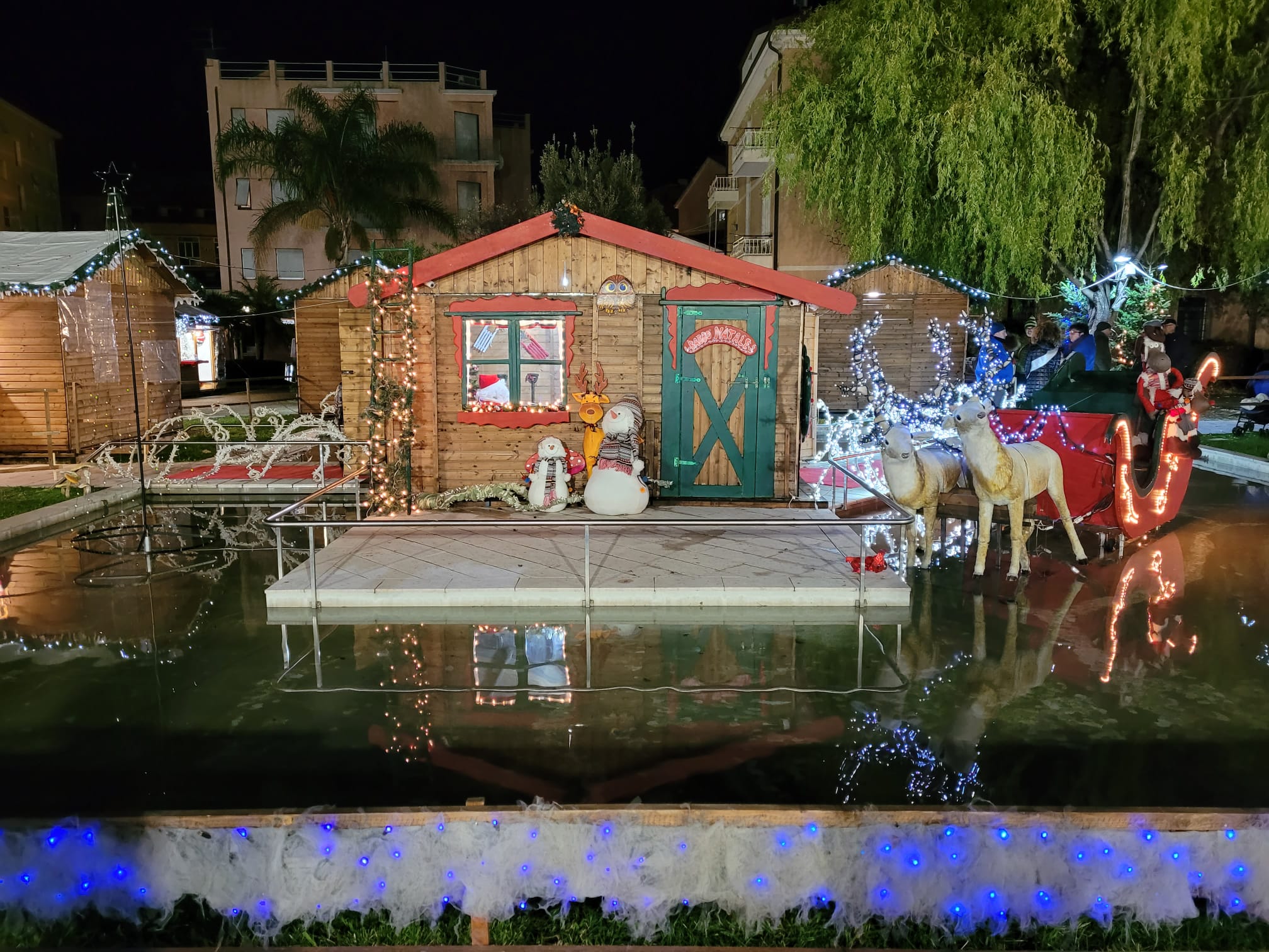 Villaggio di Natale con casette basse con luci colorate e tetti rossi, davanti a una di queste, una renna dei pupazzi di neve e una slitta