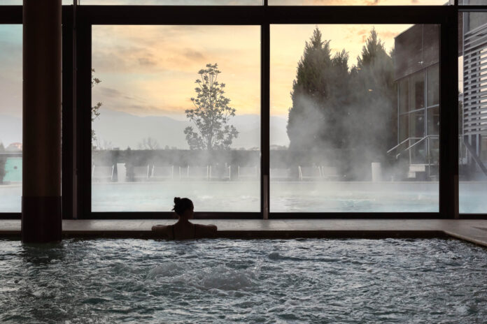 Terme Italia - la veduta di una grande piscina che affaccia su grande vetrata da cui si vedono alberi e uno splendido tramonto. Nella piscina si vede la sagoma di una donna in acqua che guarda il panorama