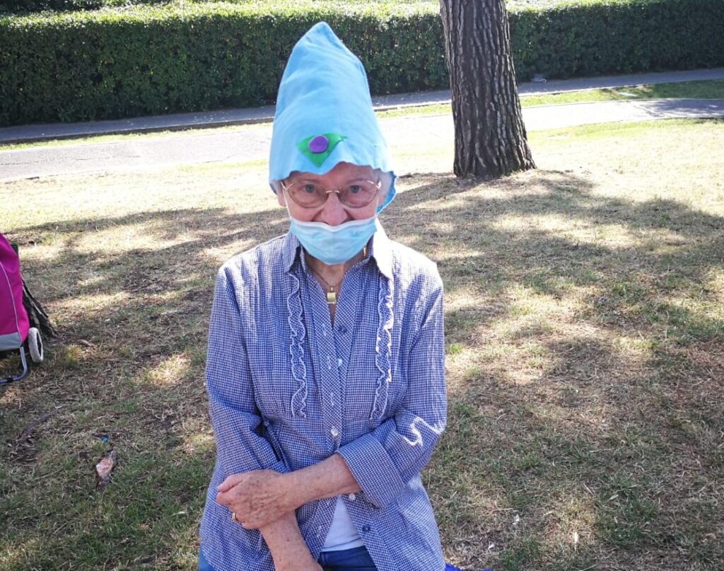 Nonna piera è una donna minuta, indossa una camicia di colore lilla con dei fiorellini, degli occhiali da vista e un cappello a cono azzuro