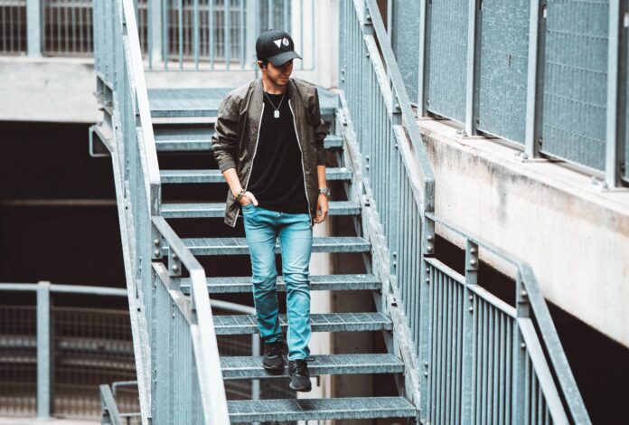 poison - MØNØ intento a scendere delle scale di metallo, indossa jeans chiari, t shirt nera, giubbotto marrone, in testa un cappellino da baseball