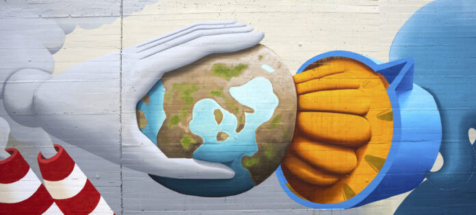 Energie rinnovabili - un opera di streetart dove si vede una mano bianca che spreme il pianeta terra con uno spremiagrumi e dietro la mano due ciminiere bianche a astrisce rosse