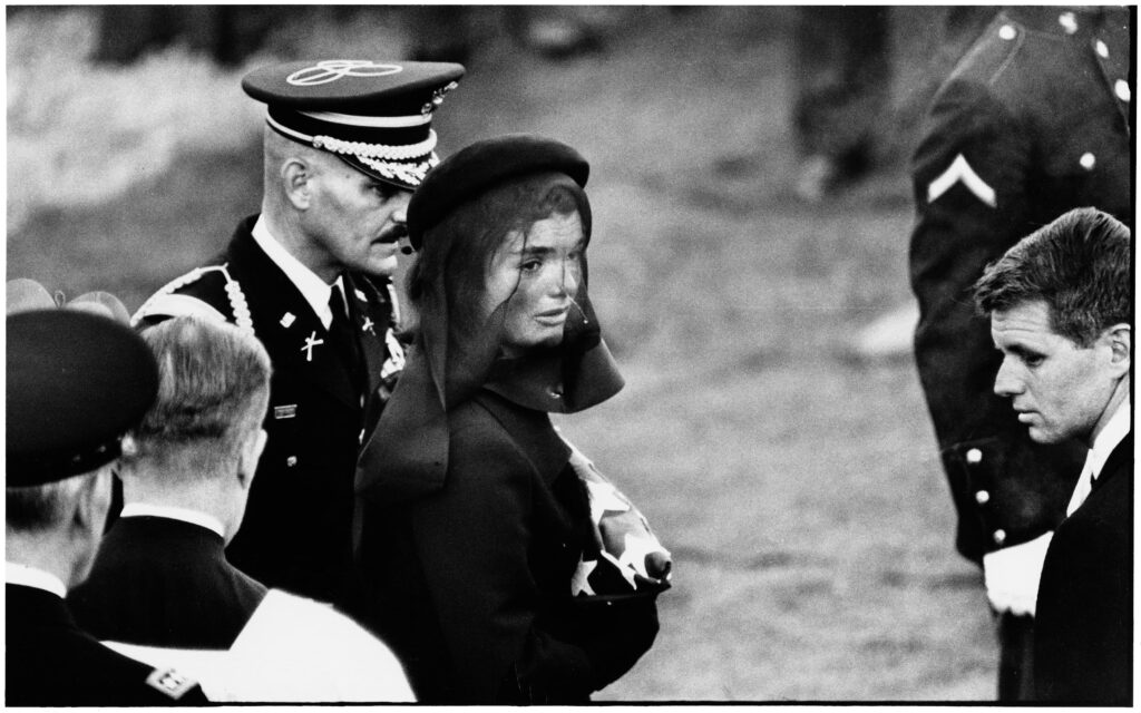 Elliott Erwitt - una foto in bianco e nero che ritrae la vedova Jaqueline Kennedy vestita di nero, al funerale del marito, vicino a lei un uomo in alta uniforme