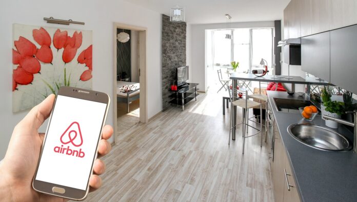 Airbnb affitti - nella foto un appartamento arredato moderno con una cucina sulla destra, un tavolo e al fondo una porta finestra. In prmo piano un amano tiene un cellulare con la schermata fissa sul logo di Airbnb