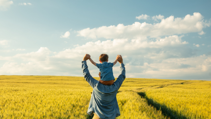 un uomo tiene sulle proprie spalle un bambino con di fronte un grnde campo di grano giallo sotto un cielo azzurro
