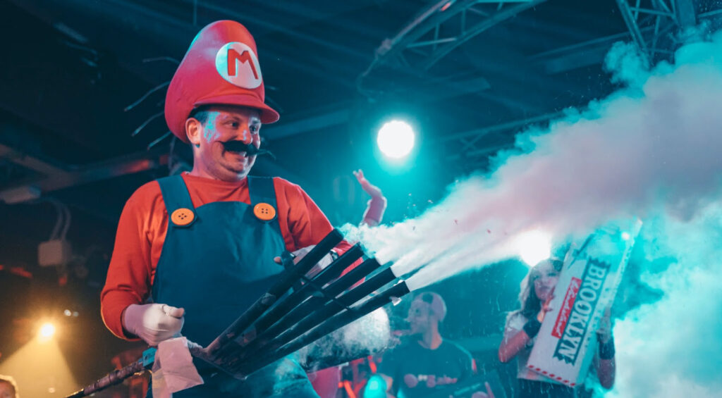 Treviso - Un uomo vestito da mario bros con cappello rosso con la lettera m, tuta blu da idraulico tiene una specie di drante con tante canne e annaffia di fumo bianco la gente che balla