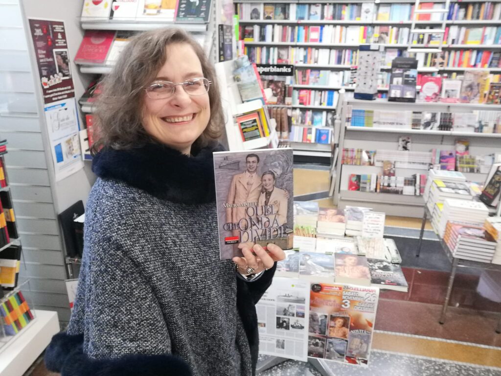 maura mafferi in piedi con capotto daavnti a uno stand interno di una libreria con il romnzo quel che onda divide in mano