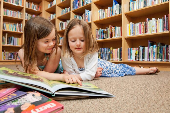 Fondazione Compagnia San Paolo premia Sbam - nella foto due bambini sdraiate per terra in una grande biblioteca leggono dun libro illustrato, dietro di loro grandi scaffali pieni di libri