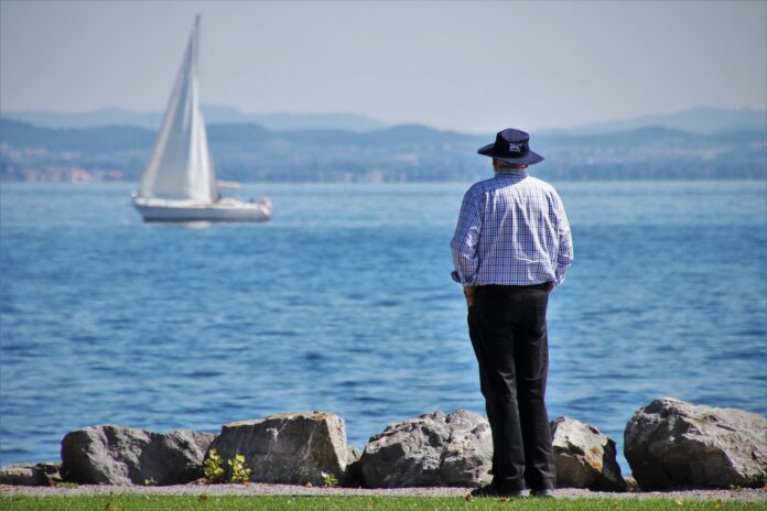 silver welfare - un anziano con il cappello, camicia binca e blu, con le mani in tasca, è sulla riva del mare e guarda una barca a vela
