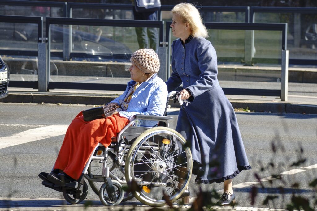 pensionati - una badante vestita con un lungo soprabito azzurro spinge una carrozzella con una signora anziana seduta. L'anziana donna indossa un cappello a bunnet di lana beige, una camicia azzurra e dei pantaloni rossi