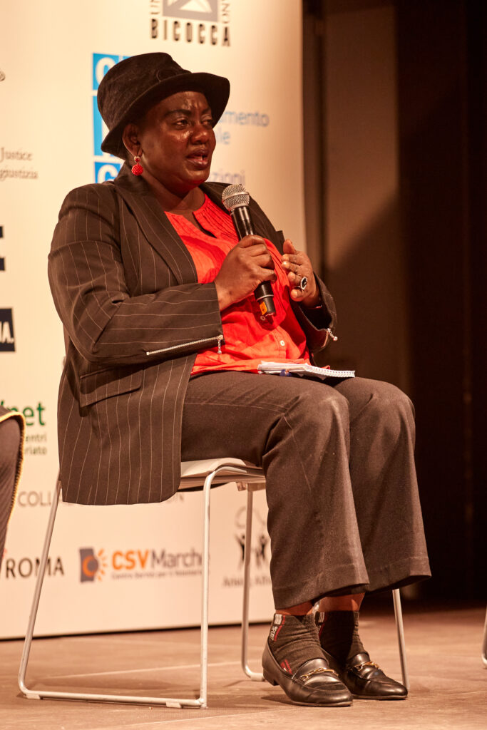 una donna nera seduta durante un convegno MGF con panaloni marroni cappello e camicia fucsia