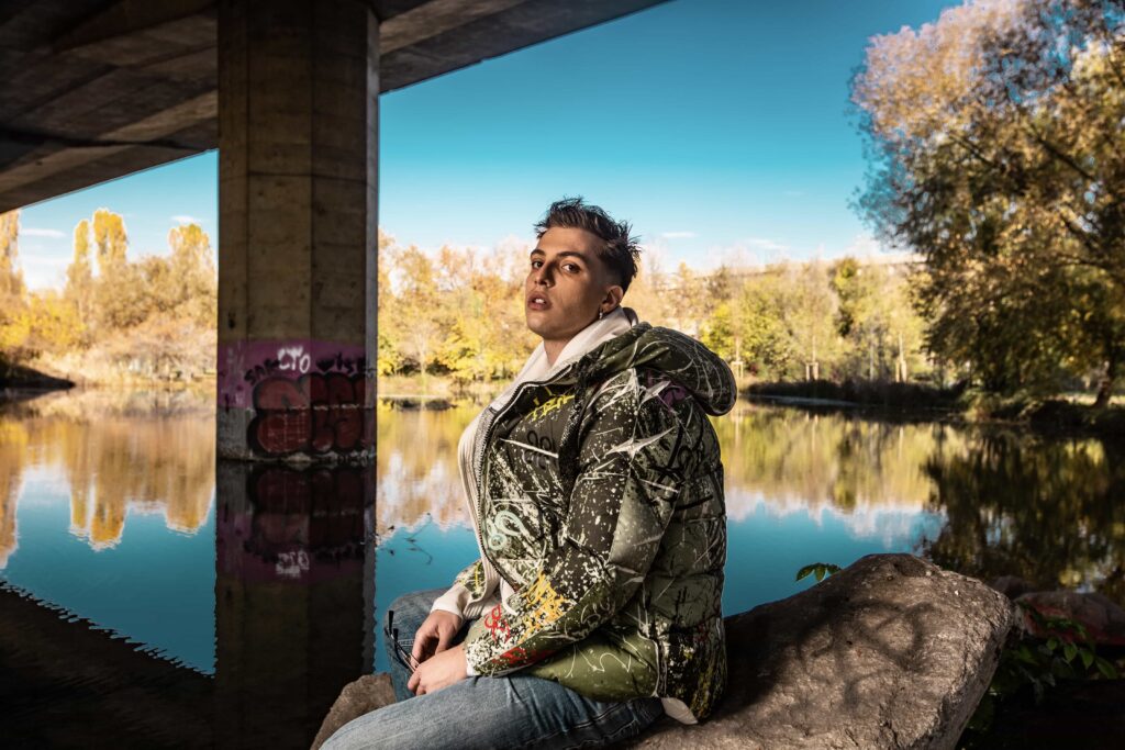 Fisso - il giovane rapper indossa jeans e una giacca a vento colorata, è seduto sotto un ponte, alle sue spalle un laghetto