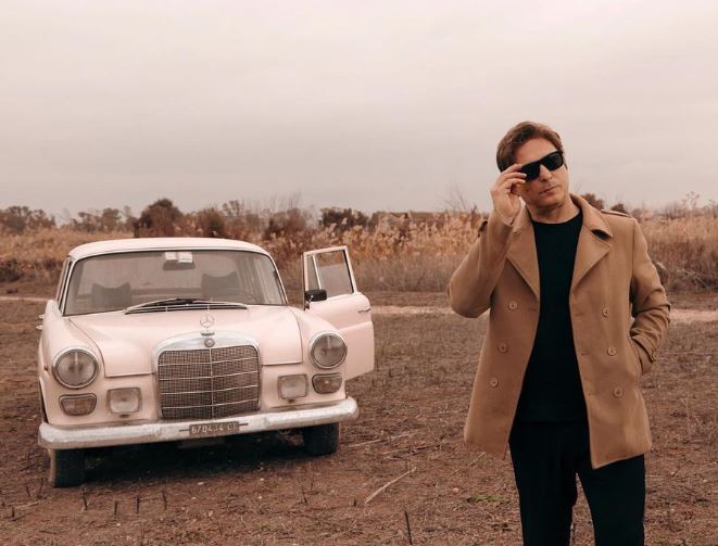 giuseppe cucè in primo piano indossa un vestito nero e un cappotto beige. sullo sfondo una nercedes bianca d'epoca