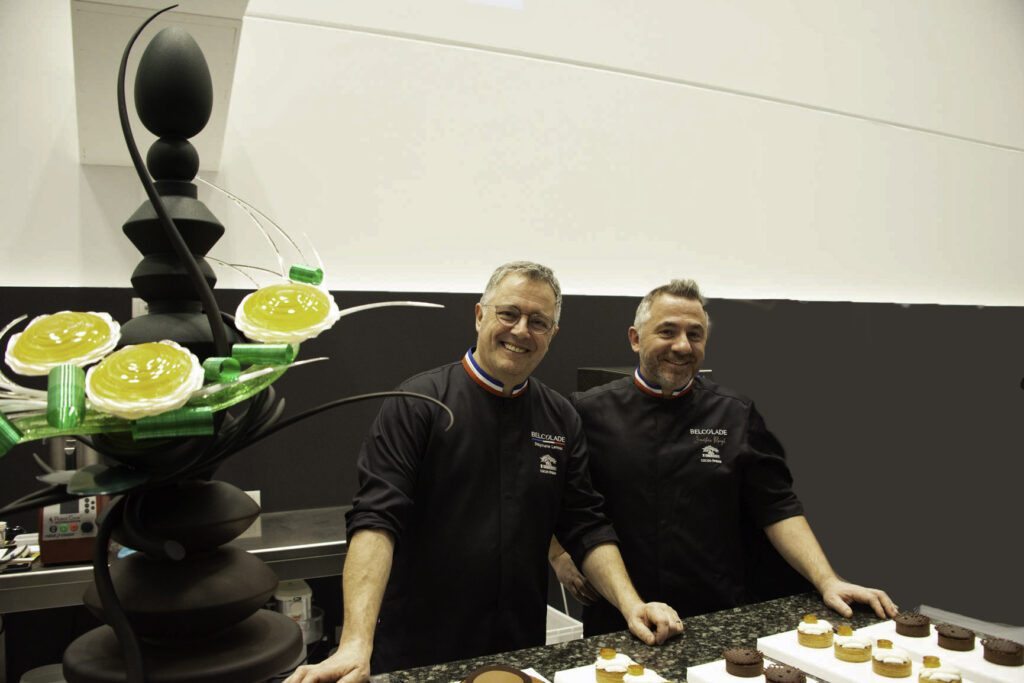 cioccolato pasticceria e arte bianca _ nella foto i due maitre chocolatier con casacca nera, sorridono, con le pmani appoggiati al banco di cucina. In primo piano, la piece di cioccolato formata da tanti piani di ovali di cioccolato che da marrone piano piano diventa nero. Al centro una scultura di zucchero fatta con tre girasoli giallli