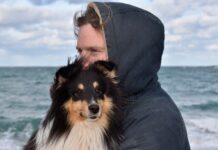 Furto di cani - nella foto un cane a pelo lungo di piccola taglia, con la testa nera e il muso marrone, è in braccio al suo padrone che indossa una felpa blu con cappuccio e dietro di loro il mare