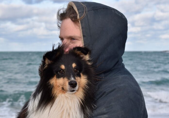 Furto di cani - nella foto un cane a pelo lungo di piccola taglia, con la testa nera e il muso marrone, è in braccio al suo padrone che indossa una felpa blu con cappuccio e dietro di loro il mare
