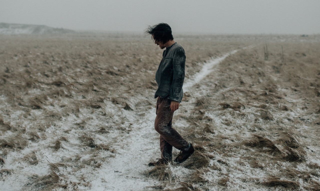 damien rice all'aperto, in un campo coperto di fango, indossa pantaloni marroni e camicia azzurra