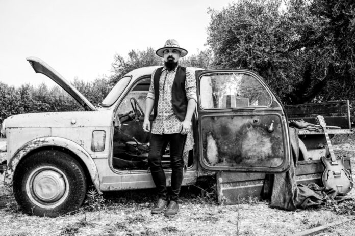 bonny jack è in piedi davanti alla carcassa di una vecchia auto, indossa cappello da cowboy, jeans neri, camicia chiara e gilet nero