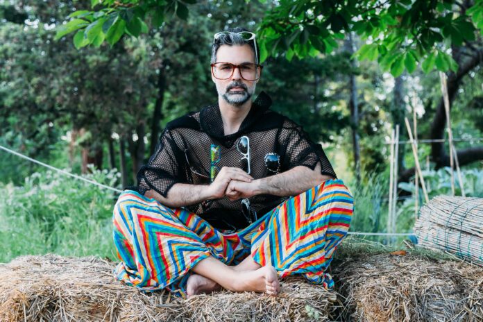 giosef, barba brizzolata e occhiali da vista, è seduto sopra una balla di fieno, indossa pantaloni a righe diagonali colorate e una camicia a rete di colore nero. ha gambe e braccia incrociate