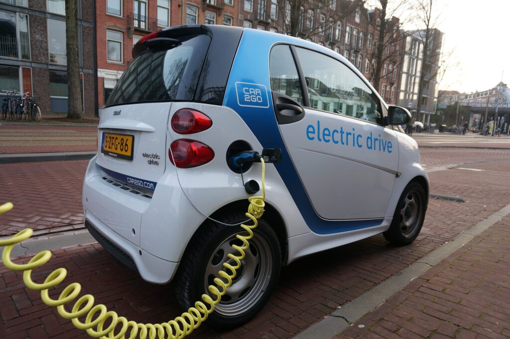 auto elettrche in car sharing - nella foto l'auto è bianca cn strisce azzurre ed è attaccata alla colonnina di ricarica tramite un cavo giallo a spirale