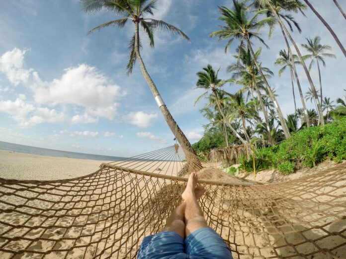 Vacanza - nella foto in primo piano un'amaca su cui si vedono le gambe di un uomo sdraiato, e una magniica spiaggia caraibica