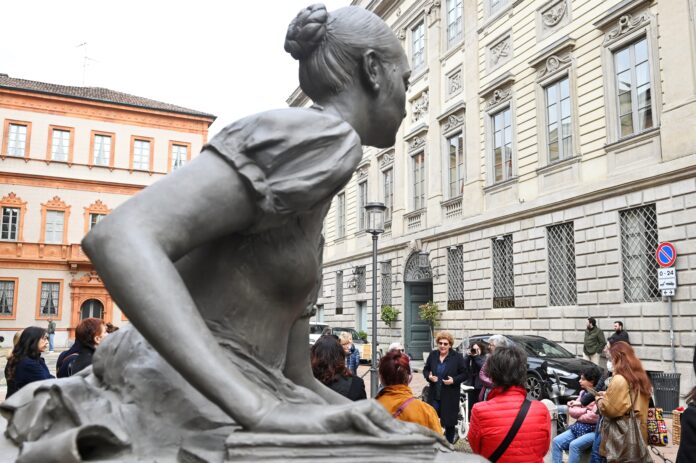 camminata in MIlano di tante donne e in primo piano la statua di un adonna con capelli raccolti dietro la nuca, inginocchiata