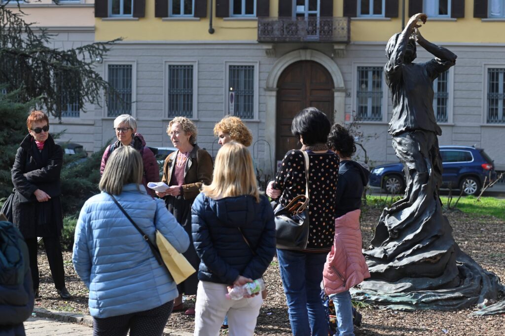 delle donne disposte in cerchio in un parco, ascoltano una guida che speiega chi era la donna rappresentata dalla statua davanti a loro