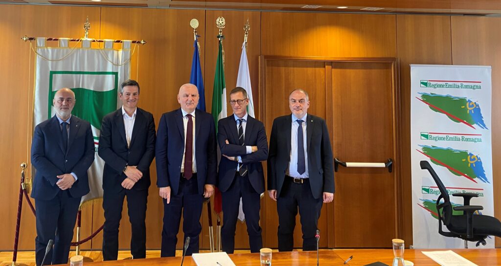 energia autoprodotta - 5 uomini vestiti tutti con giacca scura e camicia bianca, all'interno dell'aula del Comune con pareti e porta in legno, affianco a loro un pannello con il disegno della regione dell'Emilia Romagna