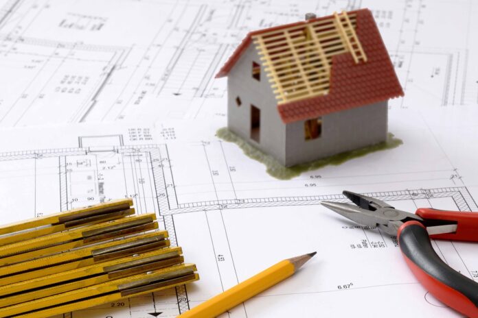 ristrutturazioni edilizia - il modellino di una casetta con il tetto rosso appoggiata sulla planimetria di un acasa, con un metro da muratore e una pinza da elettricista rossa e una matita gialla