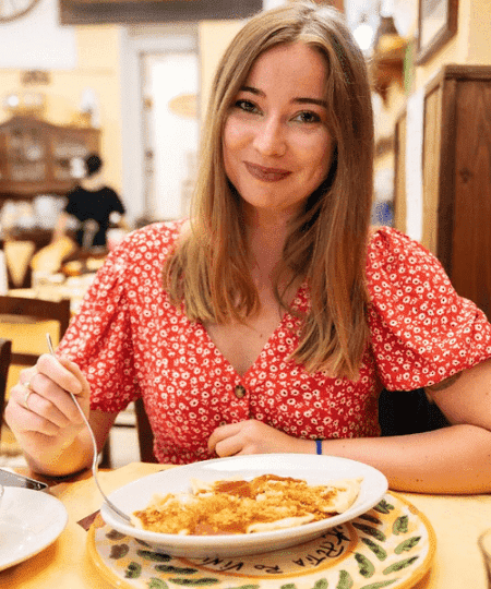 turismo in sicilia - una donna giovane e bionda sorride seduta a tavola davanto a un piatto di pasta. Indossa un vestito estivo rosso con dei fiorellini bianchi