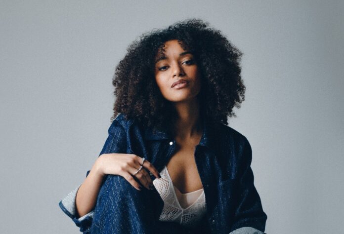 Kaze - bellissima ragazza afro americana con lunghi capelli neri e ricci è seduta e indossa un giubbotto di jeans e sotto un corpetto bianco