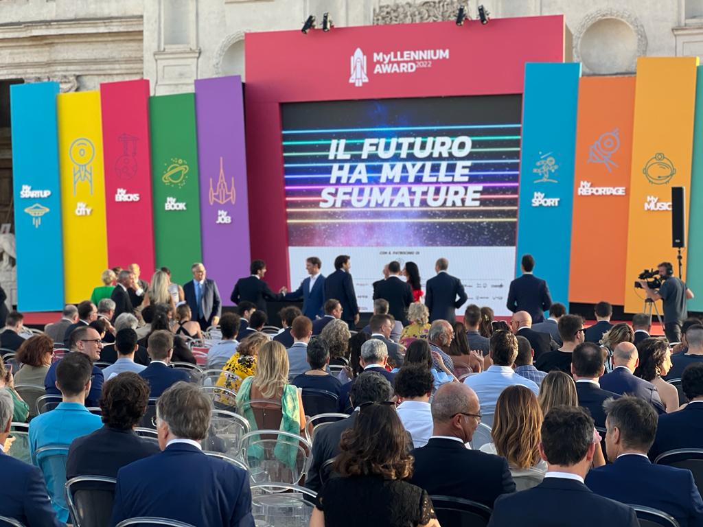 Myllennium Award mobilità sostenible - delle persone sedute ascoltano gli oratori che sono sul palco che è formato da un grande schermo con dei pannelli verticali colorati di viola, verde, rosso e viola e delle scritte appaiono su un grande schermo