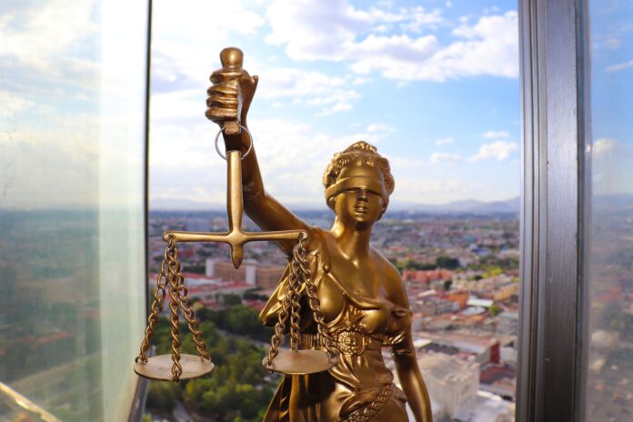 avvocati - nella foto una statua dorata della dea della giustizia bendata che tiene in mano la bilancia. La stauta è posizionata davanti a una finestra dalla quale si vede una vista panoramica di una città