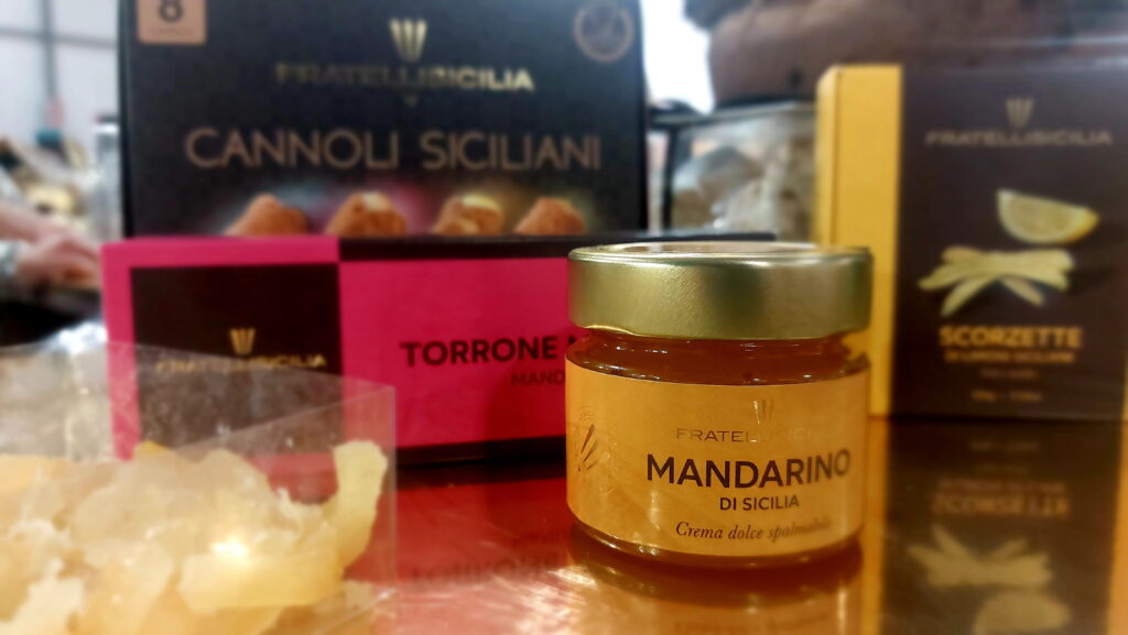 Sicilia - un barattolo di crema al mandarino con coperchio dorato e dietro una confezionemarrone con dei cannoli siciliani. In primo piano anche un vasetto con delle bucce candite