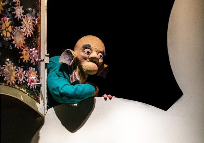 una marionetta si affaccia da una finestra nella scenografia di un teatro