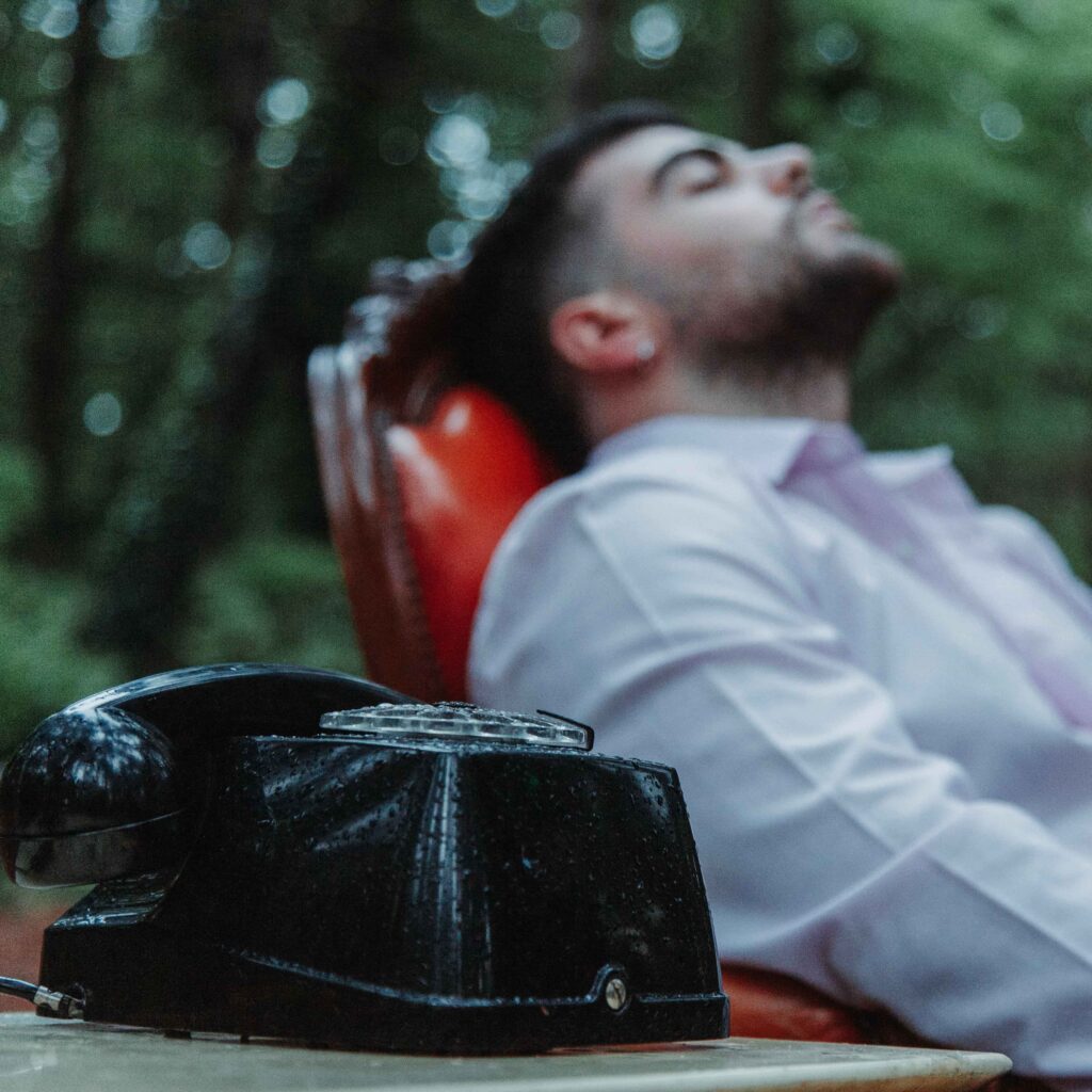 gala è seduto su una poltrona di legno e pelle rossa, indossa una camicia bianca e di fianco ha un telefono nero stile anni 60