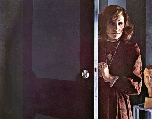 mia martini appoggiata ad una porta socchiusa, la mano sinistra sulla maniglia, ha i capelli lunghi e indossa un lungo vestito marrone