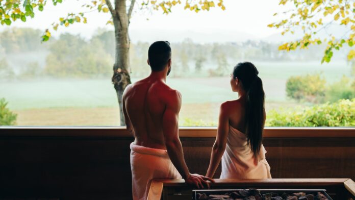 Monticello SPA & FIT ambiente - un uomo e una donna stanno guardando un giardino con alberi da un'enorme finestra panoramica della sauna