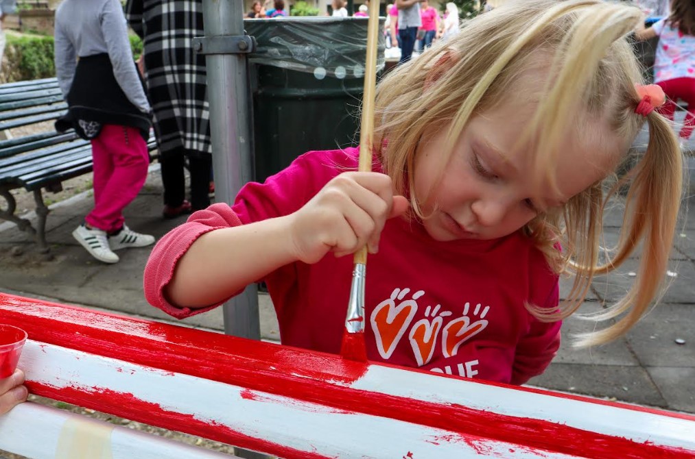 una bambina molto piccola bionda con maglia rosa pittura di rosso la panchina delle famiglie per progetto Torino Rosso indelebile