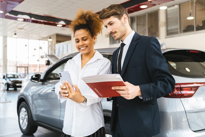 Un uomo e una donna stanno parlando sorridendo durante la trattativa di un acquisto di un auto. L'uomo tiene in mano una cartellina rossa e indossa giacca scura, camicia bianca e cravatta scura. La donna, di colore, ha capelli ricci legati e indossa una camicetta bianca. Dietro di loro un auto grigia