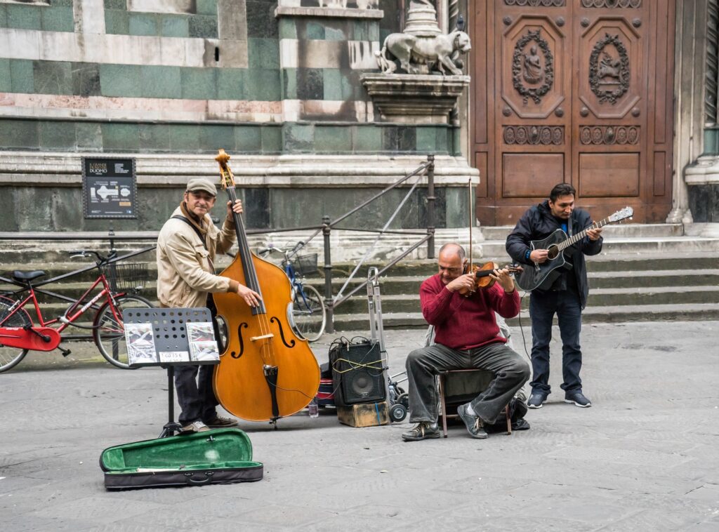 musicisti di strada, uno suona il contrabasso, uno il violino e un altro la chitarra. Per terra ci sono degli amplificatori e la custodi della chitarra aperta. Dietro di loro dei gradini di un edificio