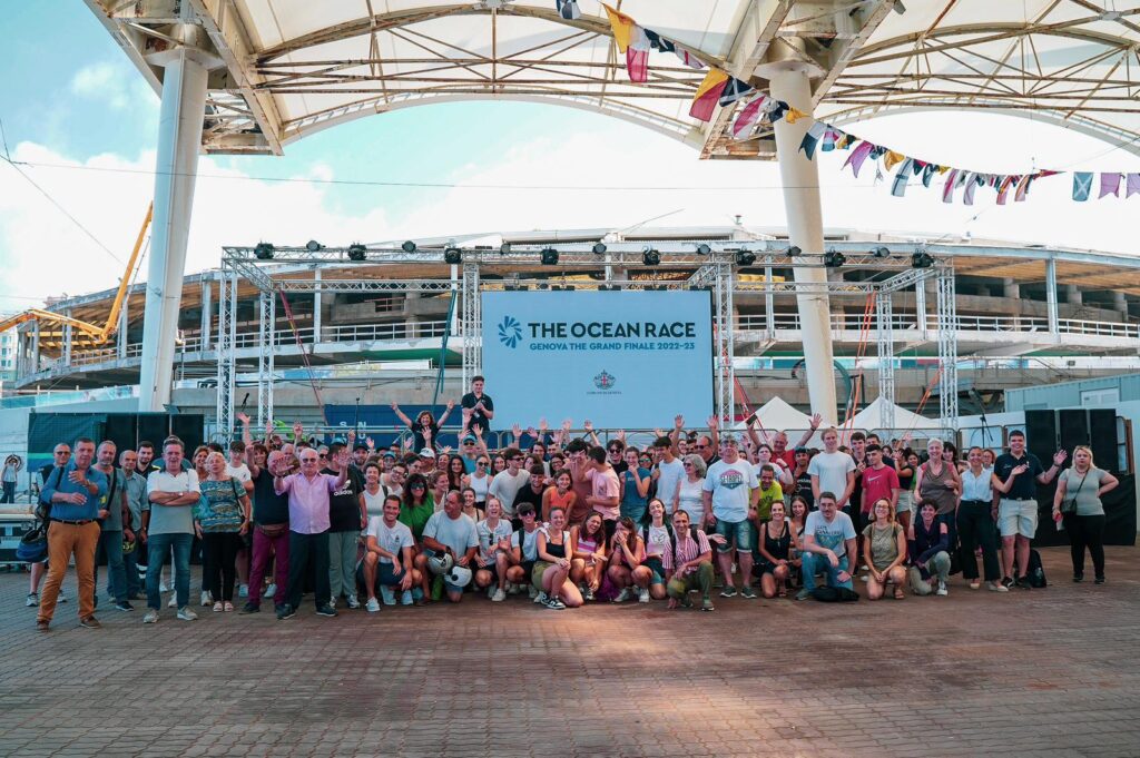 tante persone che posano per una foto di gruppo e dietro di loro un cartellone azzurro con la scritta "The Ocean Race"