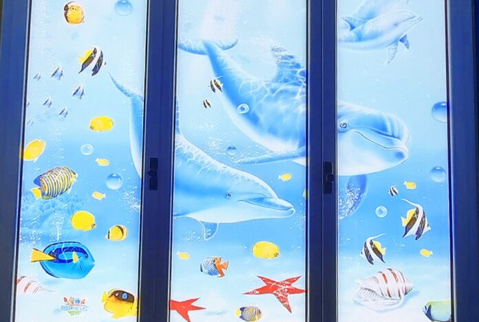 I-colori-del-mare-al-Gaslini-con-Ospedali-Dipinti - una finestra a tre ante con disegnati sui vetri dei delfini, delle stelle marine e tanti pesciolini gialli con strisce nere
