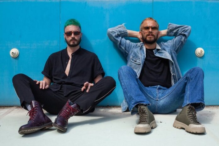 management - i due componenti la band seduti a terra, appoggiati a un muro dipinto di azzurro