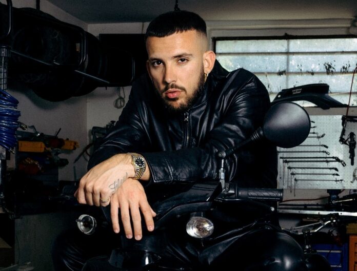 vegas jones, capelli e barba scuri, indossa un giubbotto di pelle e ha le braccia appoggiate sul manubrio di una moto